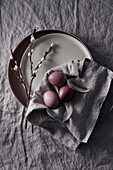 Purple Easter eggs with feathers on a grey linen cloth, willow catkins on a plate