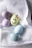 Pastel-coloured Easter eggs and yellow Easter egg with painted smiling face and flowers on white cloth