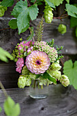 Kleiner Herbststrauß mit Dahlien (Dahlia) und Hopfen (Humulus) in Glasvase an Holzwand