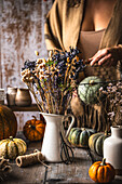 Getrocknete Blumen in weißer Vase auf Holztisch mit Kürbissen und Bastelmaterial, Herbstdeko