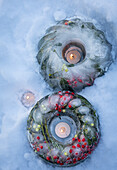 Two frozen wreaths with holly (Ilex), mistletoe and candles in the snow as Christmas decorations