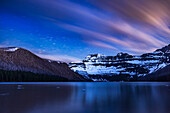 Die Sterne und Wolken ziehen am Himmel über dem Cameron Lake im Waterton Lakes National Park in Alberta und dem Mt. Custer im Glacier National Park in Montana vorbei. Der Himmel und die Berge werden von einer zunehmenden Mondsichel rechts im Bild beleuchtet, die im Nordwesten untergeht. Die Sterne des Skorpions gehen links im Bild auf. In diesem Alpensee ist noch etwas Eis vorhanden, wie Anfang Juni 2024. Durch den Wind und die lange Belichtungszeit sind die Oberfläche des Sees und die Spiegelungen verschwommen. Der Hügel links ist durch den Waldbrand von 2017 vernarbt.