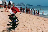 A makeshift small traditional Christmas tree and Santa Claus hat planted on the sand at Leblon Beach with a defocused crowd in the background, Rio de Janeiro, Brazil, South America