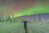 Rückansicht eines Mannes mit ausgestreckten Armen in der Mitte einer leeren, glatten Straße, die den arktischen Wald durchquert, während er die Nordlichter (Aurora Borealis) bewundert, Finnisch-Lappland, Finnland, Europa