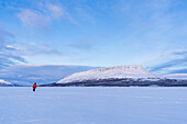 Mann in arktischer Landschaft spaziert auf der eisigen Oberfläche eines finnischen Sees vor dem Saana-Hügel (Fjäll) in der Abenddämmerung, Kilpisjarvi, Gemeinde Enontekio, Finnisch-Lappland, Finnland, Skandinavien, Europa