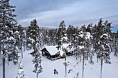 Drone view of a cyclist with fat bike standing in the middle of the tall-tree coniferous forest covered with snow in front of a mountain hut, Finnish Lapland, Finland, Scandinavia, Europe