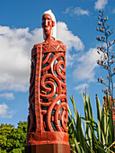 Schnitzerei einer Figur in einem Flachsmantel neben einer Flachspflanze in Te Puia, Gisborne District, Nordinsel, Neuseeland, Pazifik