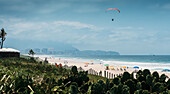 A paraglider soars above a tranquil beach dotted with colorful umbrellas, with a backdrop of a clear blue sky in Barra da Tijuca, Rio de Janeiro, Brazil, South America