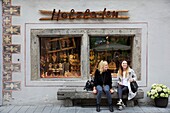 Italy, autonomous province of Bolzano, Brunico, two women sitting on a stone bench in front of a craft shop in a downtown alley