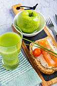 Top view of a nutritious meal with a slice of whole grain toast topped with smoked salmon, cream cheese, and a cherry tomato, accompanied by a fresh green apple and cucumber juice. Served on a wooden board with a modern, marble background.