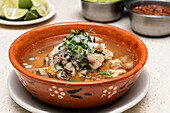 Authentic Mexican pozole, a traditional soup featuring hominy and pork, garnished with chopped onions and cilantro, presented in a rustic clay bowl with carved designs. Lime wedges and salsa accompany this hearty dish.