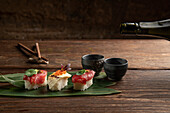 An elegant presentation of sushi delicacies, laid on a bamboo leaf, accompanied by sake cups, on a rustic wooden table setting, ideal for a sophisticated dining experience