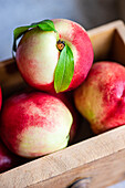 A close-up image of vibrant, seasonal nectarines with a leaf on, set in a rustic wooden crate, highlighting their fresh and juicy appearance