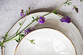 Draufsicht auf einen minimalistisch gedeckten Sommertisch mit einem Vintage-Teller und wilden Alfalfa-Blumen, die einen Hauch von natürlicher Eleganz vermitteln.