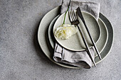 A tasteful table setting featuring stacks of gray ceramic plates, a silver fork and knife, and a delicate white Godetia flower, presented on a textured grey backdrop