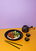 Artfully plated teriyaki chicken on a black plate, served with broccoli and rice, accompanied by a traditional tea set on a vibrant yellow and purple background.