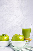 Fresh green apples in a white bowl next to a glass of nutritious cucumber and apple juice, elegantly placed on a marble surface. The scene is complemented by a soft green napkin, emphasizing a healthy lifestyle.