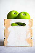 Ripe green apples are neatly placed in a rustic wooden crate with a bright white backdrop, highlighting their vibrant color and freshness