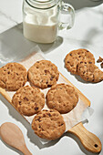 Delicious chocolate chip cookies on a baking paper beside a glass jar of milk and a wooden spoon, captured from the top view under natural lighting