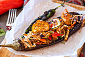 Baked stuffed eggplant with cherry tomatoes, bell peppers, onion, garlic, thin potato slices, cheese, and microgreens served on parchment paper."