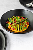 A vibrant dish of mashed sweet potato garnished with grilled asparagus, mint leaves, and pomegranate seeds in a black dish, shot from an angled view.