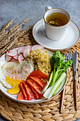 Ein appetitlicher Teller mit geschnittenem Rettich, Tomate, Sellerie, Spiegelei und Bulgur-Getreide, dazu eine Tasse heißer Tee, auf einer Weidenmatte.