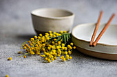 A serene table setting featuring ceramic tableware with copper chopsticks and a scattering of bright mimosa flowers adding a pop of color