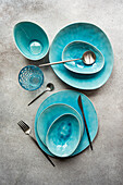 Top view of a stylish turquoise ceramic table setting with matching cutlery on a textured surface.