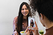 Unrecognizable African American female taking photo of friend sitting at table with poke dish in restaurant