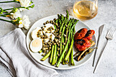 A nutritious meal featuring a boiled egg, BBQ asparagus, capers, and grilled vegetables including tomatoes and chili peppers, presented stylishly on a ceramic plate.