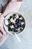 A nourishing bowl of cottage cheese and sour cream topped with organic blueberry jam and fresh blueberries, served with a spoon on a light gray surface, accompanied by a pink linen