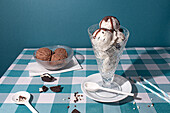 A tantalizing ice cream sundae drizzled with syrup alongside chocolate cookies in a casual table setting with a blue backdrop