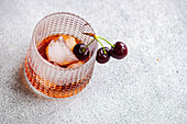 A sophisticated glass of cherry liqueur served on ice, garnished with a pair of ripe cherries, presented on a textured surface