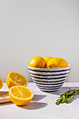 A ceramic bowl with vertical stripes filled with bright yellow lemons, with halved fruits and fresh herbs on a light surface