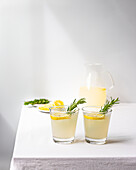 Two glasses of homemade lemonade with rosemary sprigs and a pitcher in the background on a white table, providing a fresh and clean look