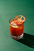 A well-prepared Bloody Mary cocktail garnished with lemon and dried tomato on a green backdrop, casting a sharp shadow.