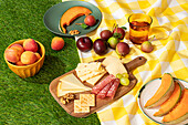 A vibrant summer picnic display featuring a selection of fruits, cheeses, and meats arranged neatly on a yellow and white checkered blanket laid on artificial grass