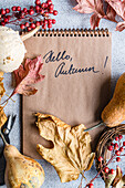 Craft styled autumnal flat lay featuring natural elements like dried leaves and gourds, a notebook with HELLO, AUTUMN message, and seasonal decorations