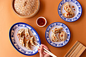 A vibrant image showcasing steamed Chinese dumplings, drizzled with spicy sauce and sesame seeds, elegantly plated on blue and white porcelain dishes against a warm orange background