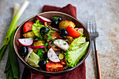 Close-up of a hearty bowl of Mediterranean salad, garnished with dill, featuring roasted potatoes, olives, cherry tomatoes, spring onions, crisp lettuce, and fresh radishes