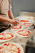 Ein anonymer Koch bereitet in einer Großküche Pizzen zu, indem er frischen Teig mit Soße und Käse belegt Bild mit Fokus auf Hände und Pizzen, mit gesichtslosem Ausschnitt