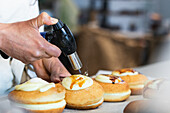 Unrecognizable crop chef caramelizing sugar cream on sweet Berliners with gas torch while preparing vegan dessert in bakehouse