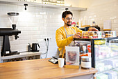 A smiling male barista in a yellow sweater is arranging a selection of pastries in a display case in a contemporary café.