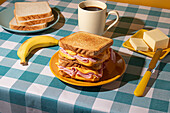 Sandwich on a yellow plate accompanies a cup of coffee, butter on a small plate, and a fresh banana, all arranged on a blue and white checkered blue tablecloth