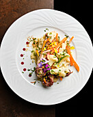 A meticulously prepared gourmet dish features seafood, julienne carrots, and colorful edible flowers, artistically plated on a white dish against a dark background