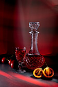 A sophisticated display featuring a crystal decanter filled with red liquid, accompanied by a matching glass, fresh cherries, and halved tamarillo on a dramatic red backdrop