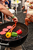 Cropped unrecognizable person grilling sausages and vegetables on a barbecue at an outdoor gathering. Background includes guests socializing, enjoying drinks and music.