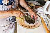 Eine ältere Frau mit Schürze ist in ihrer Küche zu sehen, wie sie über einem Teller eine Aubergine schält, wobei der Fokus auf ihren Händen und dem Gemüse liegt