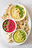 Top view of colorful assortment of classic, beet, and avocado hummus, each garnished and served with pita bread, showcasing vibrant, healthy dips for snacking or appetizers.