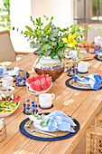 A vibrant and colorful breakfast set on a wooden terrace table, featuring a lush floral centerpiece and a selection of fresh fruits